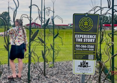 A person interacts with a metal sculpture garden outdoors. In the foreground, a sign invites visitors to “Experience the Story” via phone or app for the Talking Trail, with contact details and a QR code.