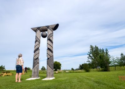 A person stands on green grass looking up at a tall wooden sculpture with two textured pillars and a suspended black sphere, under a partly cloudy sky in an open park.