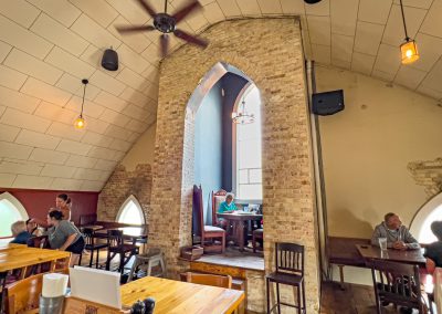 A cozy café with vaulted ceilings, brick archways, and gothic-style windows. Several groups of people are seated at wooden tables, chatting and enjoying their time in the warmly lit space.