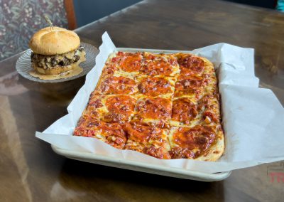 A rectangular pepperoni pizza cut into squares sits on a tray lined with parchment paper next to a plate with a burger topped with mushrooms and cheese, both placed on a wooden table.