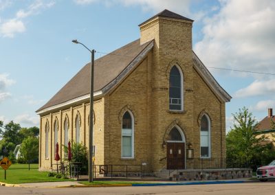 A historic yellow-brick church with arched windows stands on a corner lot, surrounded by grass and trees, with a streetlamp and parked cars nearby under a partly cloudy sky.