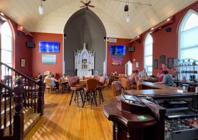 A spacious bar set inside a former church, with high arched ceilings, stained glass windows, a central altar-like structure, and people seated at tables and the bar, enjoying drinks.