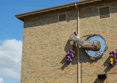 A large art installation of a mirrored circle with wooden logs and purple geometric shapes is mounted on a tan brick building under a blue sky with clouds.