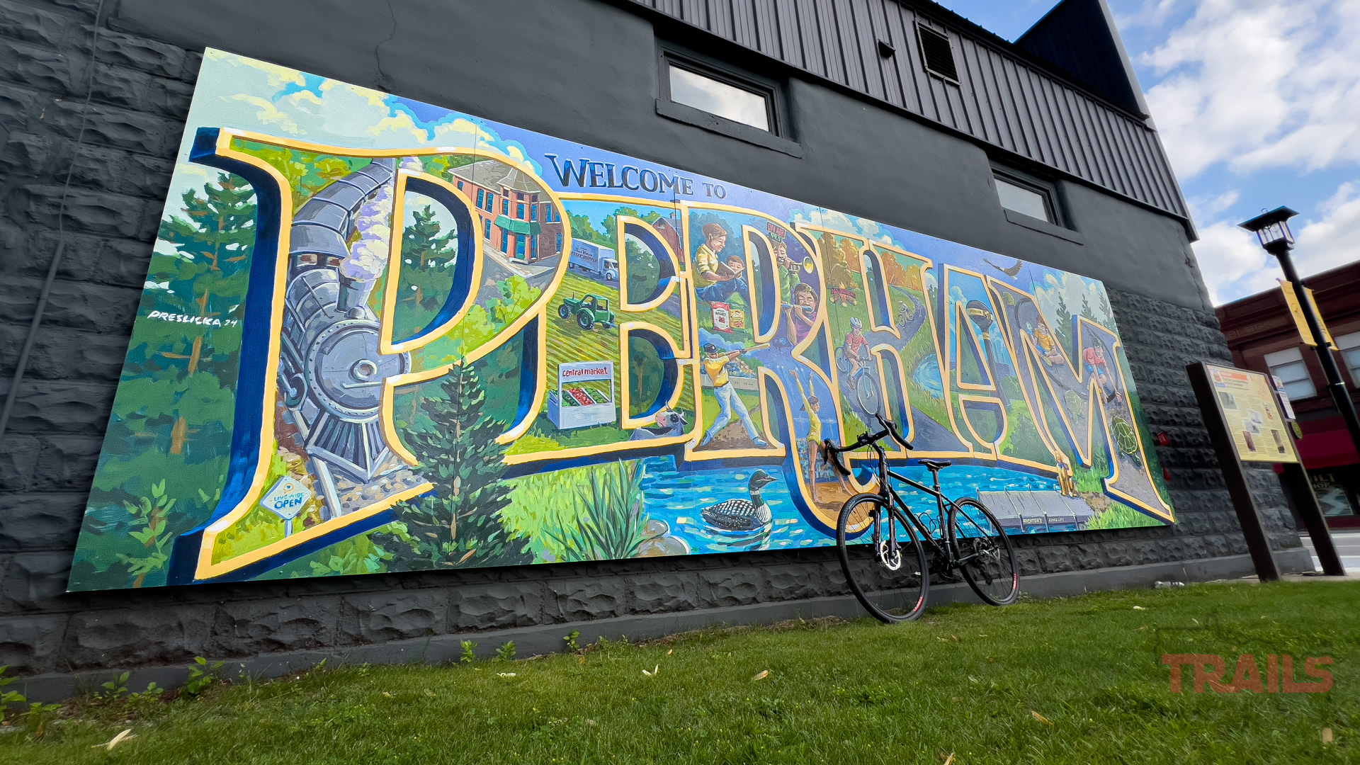 Perham Bicycling A colorful mural on a black brick wall reads Welcome to Perham with local scenery inside bold letters. A bicycle is parked in front, and the sky is partly cloudy.