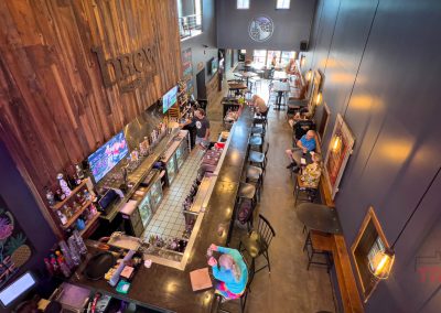 A view from above of a modern bar with a long counter, several people seated, wooden walls, colorful chalkboard art, and natural light coming through large front windows.