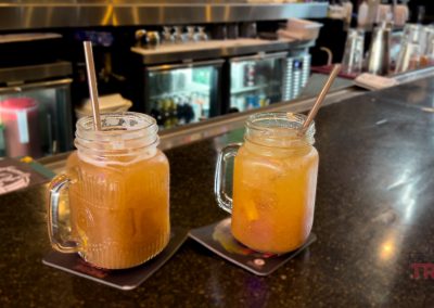 Two mason jar glasses with metal straws filled with orange-colored drinks sit on coasters on a bar counter, with a row of beverage coolers and various bottles visible in the background.