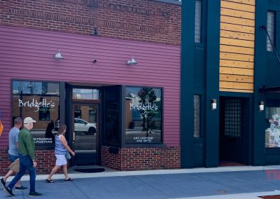 Two people walk past a storefront called “Bridgette’s Main Frames” with large display windows, a pink and red exterior, and flags hanging on the side. Reflective glass shows buildings and trees across the street.