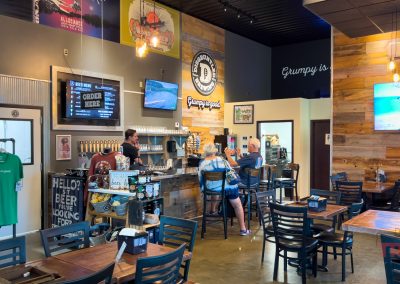 A modern brewery taproom with a bar, four people seated, wooden tables, a merchandise area with green t-shirts, wall art, and two wall-mounted TVs displaying different images; the space is warmly lit.
