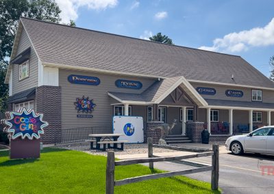 A large building with signs for popcorn, toys, games, and candy. A white car is parked in front, and a colorful Cookie Caboose sign stands near outdoor benches on a sunny day.