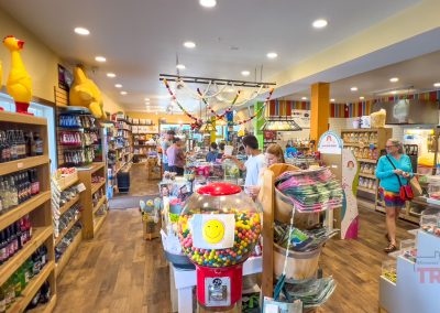A colorful candy and gift shop with shelves of sweets, snacks, and toys. Several people browse the aisles, and a large gumball machine with a smiley face stands in the foreground. Bright decor and rubber chickens are visible.