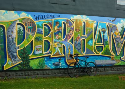 A colorful mural on a stone wall reads “Welcome to Perham” with local scenes and nature inside the letters. A bicycle is parked in front of the mural.