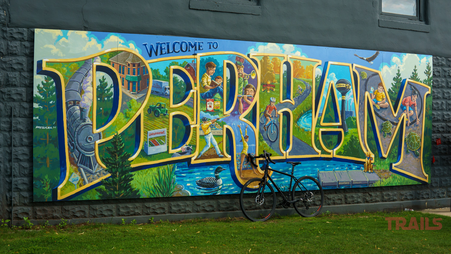 Perham-Mural A colorful mural on a stone wall reads “Welcome to Perham” with local scenes and nature inside the letters. A bicycle is parked in front of the mural.
