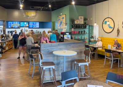 Casual café interior with wooden floors, bright lighting, and people ordering at the counter or seated at tables. Yellow booth seating runs along the right wall, and maps and mugs decorate the walls.