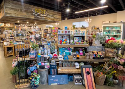 A well-lit boutique shop called Nest displays an array of colorful gifts, home decor, games, and accessories on shelves and tables. The store has a rustic wooden accent and neatly organized merchandise.