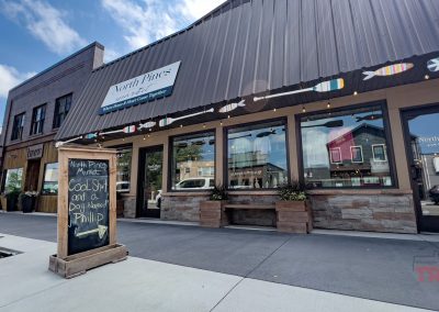 Street view of North Pines, a pet supply store with large windows and a sign featuring colorful fish. A chalkboard outside advertises “Cool sh*t and a dog named Phillip.” Other storefronts are visible nearby.