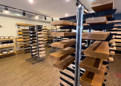 A room with several racks holding large wooden slabs displayed horizontally, likely in a wood or furniture store, with a wooden floor and track lighting overhead.