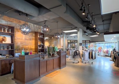 A bright, modern clothing boutique with racks of clothes, a central wooden checkout counter, and decorative lighting fixtures. Large windows let in natural light and the store has an open, spacious layout.