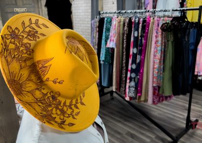 A bright yellow hat with brown floral designs is displayed in the foreground, with a clothing rack of colorful garments and patterned dresses in the background inside a boutique.