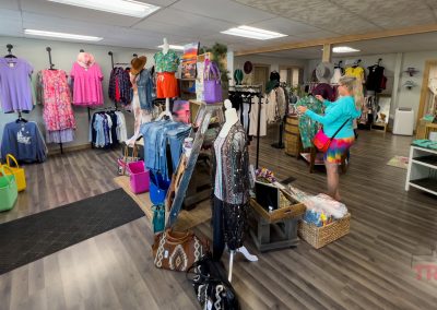 A boutique clothing store with racks of colorful dresses, shirts, and accessories. A woman in a turquoise top and rainbow shorts shops near a mannequin, and baskets and bags are displayed on the floor and tables.