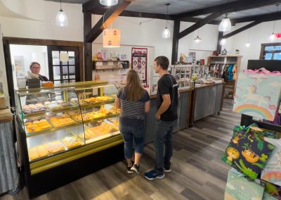 Two people stand at a bakery counter inside a shop, looking at a display case filled with donuts. A staff member is behind the counter, and shelves with gifts and colorful bags are visible around the store.