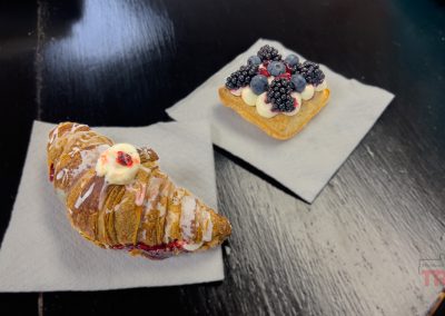 A croissant topped with icing and berries and a square tart with cream, blackberries, and blueberries sit on white napkins on a dark table.