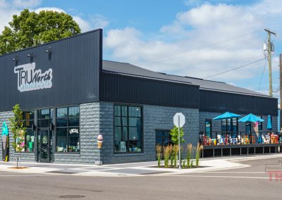 A modern black and gray building houses Tru North Minnesota, with outdoor patio seating under blue umbrellas. A clear, sunny day highlights the corner location and clean, urban surroundings.