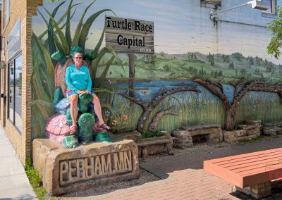 A woman sits on a colorful turtle statue in front of a mural and a sign reading Turtle Race Capital in Perham, MN. The mural depicts wetlands and reeds, with benches nearby on a brick sidewalk.