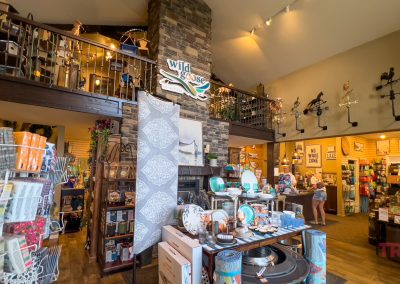 A gift shop interior with shelves of home decor, greeting cards, and other gifts. A stone fireplace, loft, and weather vanes decorate the space. Two people browse merchandise. The Wild Goose sign hangs above the fireplace.