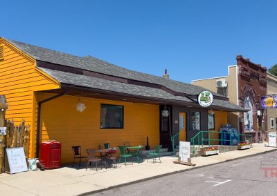 A bright yellow café with outdoor tables sits on a sunny small-town street next to older brick and tan buildings. A signpost and colorful decorations are visible near the entrance. Blue skies overhead.