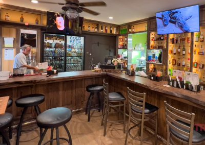 A cozy bar with wooden counters and stools, shelves of liquor bottles, a bartender behind the counter, a fridge labeled COLD BEER, and a TV showing a nature scene on the wall.