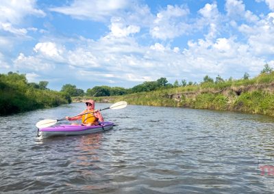 A person in an orange vest paddles a purple kayak down a narrow, calm river surrounded by green grassy banks under a blue sky with scattered clouds.
