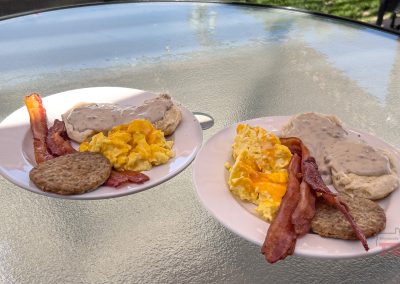 Two white plates on a glass table each hold scrambled eggs, bacon strips, sausage patties, and biscuits topped with white gravy. The scene is outdoors, with sunlight and grass visible in the background.