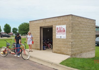 Two people stand outside a small brick building labeled Red Rock Luverne Bike Loan Program. The man adjusts a tandem bicycle, while the woman looks on. Several bikes are visible inside the open building.