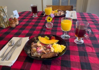 A breakfast table with a red and black checkered tablecloth, featuring plates of fruit, scrambled eggs, sausage, and pastries, with glasses of orange juice and coffee, flowers, and utensils neatly arranged.