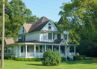 A large, white, two-story Victorian-style house with a spacious wraparound porch sits among green lawns and tall trees on a sunny day.