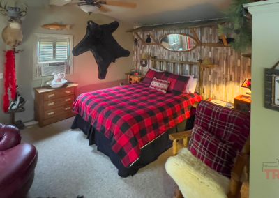 A cozy bedroom with rustic decor, featuring a bed with a red and black plaid blanket, wood-paneled accent wall, animal mounts, antlers, and a plush armchair draped with a matching plaid throw.