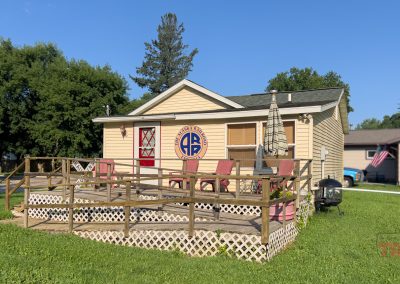 A small tan house with a wooden deck, red chairs, and an umbrella sits on a grassy lawn. The house features a round blue and yellow emblem and is surrounded by trees and neighboring homes.