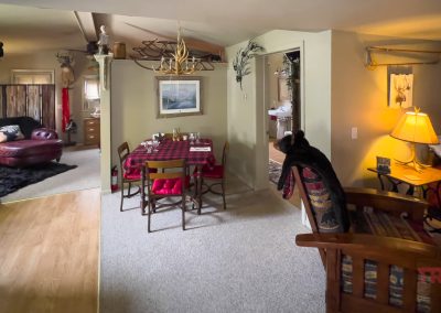 A cozy cabin interior featuring a dining table with a red plaid tablecloth, rustic wooden furniture, antler decorations, warm lighting, and a glimpse into a living and bedroom area.