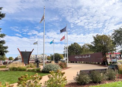 A landscaped park area in Marshall, MN, 911 Memorial Park has decorative plants, a curved metal wall with script writing, benches, under a partly cloudy sky.
