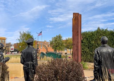 Three bronze statues—a firefighter, a police officer, and a civilian—stand facing a beam from the World Trade Center at this 9/11 memorial in Marshall, MN. An American flag and city buildings rise in the background.