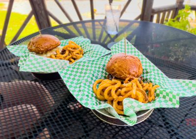 Two baskets lined with green checkered paper each holding a burger and a serving of curly fries sit on a black outdoor mesh table, with a clear drinking glass in the background.