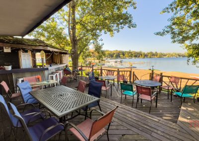 Outdoor wooden patio with colorful chairs and tables overlooking a sandy beach and calm lake; dock and boats are visible in the distance, surrounded by green trees on a sunny day.