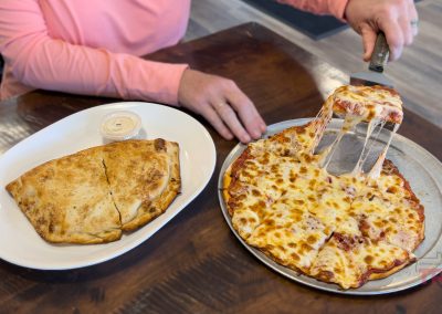 At Big John’s Pelican Pizza, a person in a pink shirt lifts a cheesy slice from a whole pizza on a metal tray, next to a calzone with dipping sauce—all served on a wooden table.