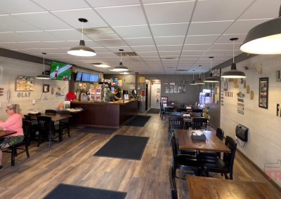 Casual restaurant interior with wooden tables and black chairs, wall art, and several hanging lights. A person sits alone at a table on the left, and the counter area with staff and TVs is visible in the background.