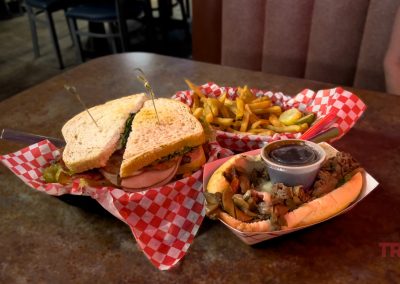 Two baskets on a table: one with a sandwich on thick bread and a side of fries, the other with a hoagie-style sandwich topped with meat and mushrooms, plus a cup of sauce. A person sits nearby wearing a floral shirt.