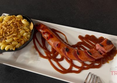 A rectangular white plate holds a grilled Currywurst drizzled with barbecue sauce and a small skillet filled with macaroni and cheese, topped with crispy onions, on a dark tabletop.
