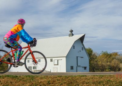 A cyclist in colorful clothing rides a red bicycle past a large white barn with a gray roof on the Camden Regional Trail, surrounded by grass and trees on a clear day.