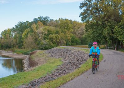 A person in a blue jacket and pink helmet rides a bicycle along the Camden Regional Trail, following a paved path beside a river, surrounded by green trees and grass under a partly cloudy sky.