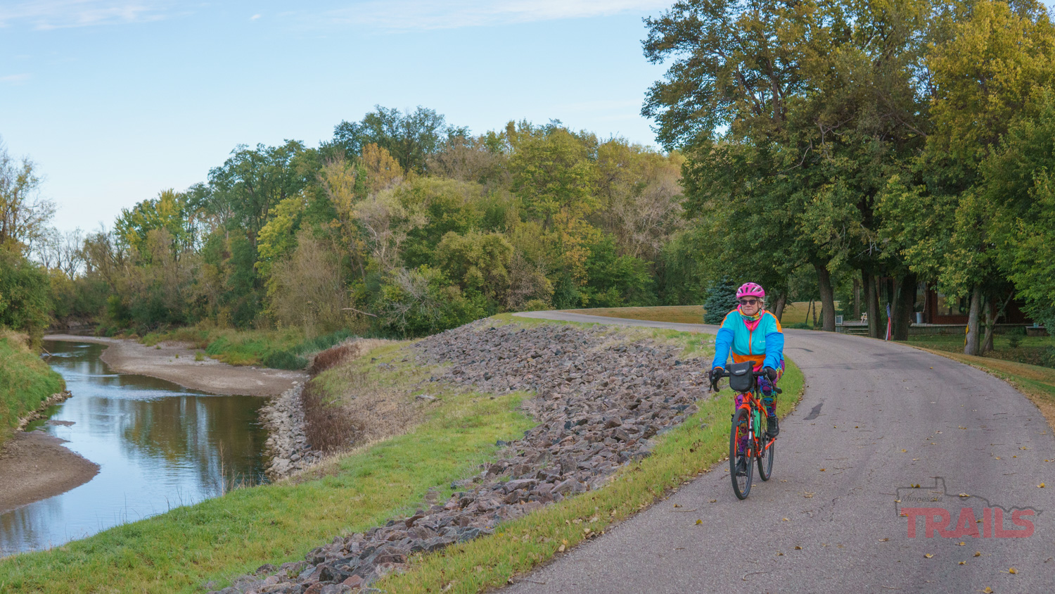 Camden Regional Trail Cottonwood River A person in a blue jacket and pink helmet rides a bicycle along the Camden Regional Trail, following a paved path beside a river, surrounded by green trees and grass under a partly cloudy sky.
