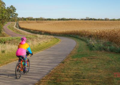 A person wearing a colorful jacket and helmet rides a bicycle on the Camden Regional Trail, a paved path that winds through fields and farmland under a clear sky.